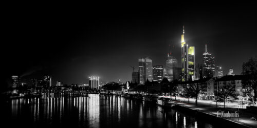 Eine Panorama Aufnahme von Frankfurt bei Nacht mit den Hochhäusern des Finanzdistrikts im Hintergrund. Schwarzweiß mit gelben und roten Elementen