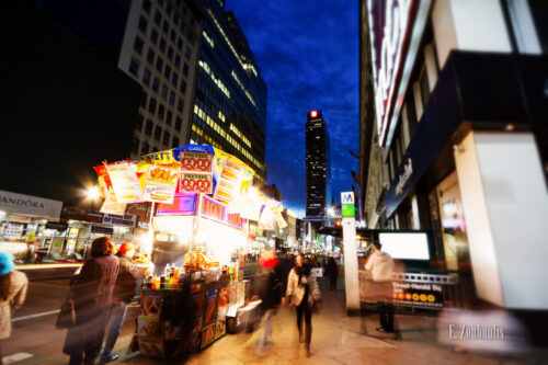 Aufnahme zur blauen Stunde in New York City. Links im Vordergrund ein Hot Dog Stand und viele Passanten, die zu jeder Zeit in NY unterwegs sind