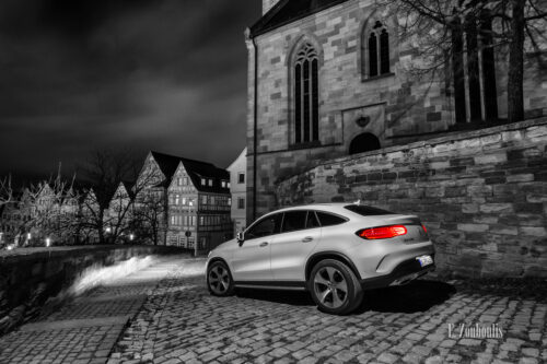 Schwarzweiß Aufnahme mit roten und blauen Elementen bei Nacht eines weißen Mercedes GLE in der Altstadt von Böblingen. Im Vordergrund das Fahrzeug auf dem Pflasterstein, im Hintergrund die Kirche und die Fachwerkhäuser am Marktplatz. Bild entstanden auf Einladung von Mercedes Benz