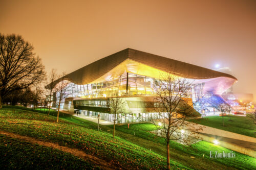 Nachtaufnahme bei Nebel an der BMW Welt in München beim Olympiapark. Im Vordergrund der Rasen, der die letzten Blätter der Bäume auffängt. In der Mitte des Bildes die BMW World mit vielen farbigen Lichtern und im Hintergrund im Nebel das Vierzylinder Gebäude