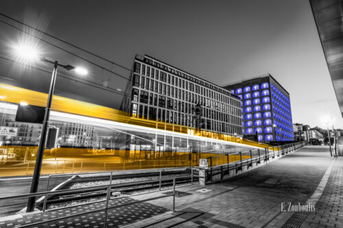 Schwarzweiß-Aufnahme an der Stadtbibliothek Stuttgart. Im Vordergrund zieht ein gelber Lichtschweif einer Stadtbahn am Betrachter in Richtung Stadtbibliothek vorbei, welche in der schönen blauen Nachtbeleuchtung zu sehen ist