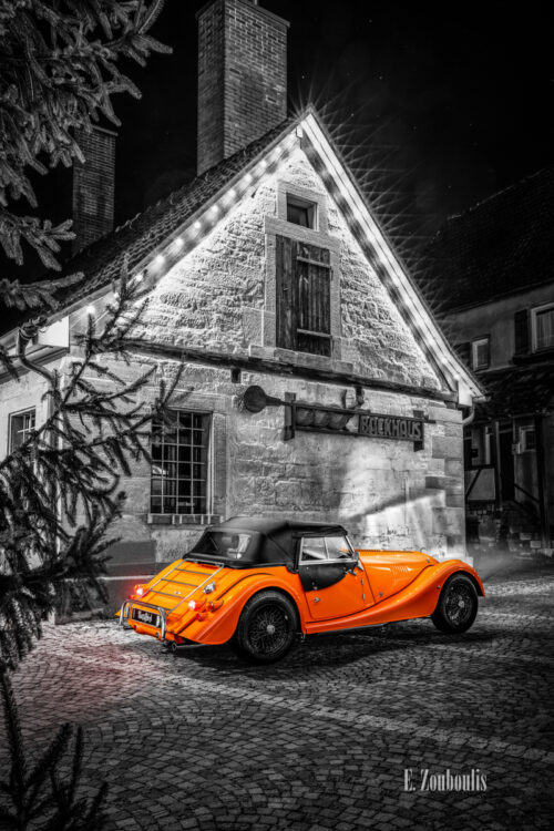 Schwarzweißaufnahme eines Orangenen Morgan 4/4. Fahrzeug in Orange in der Mitte des Bildes. Im Hintergrund ist das Backhaus am Marktplatz in Gärtringen zu sehen