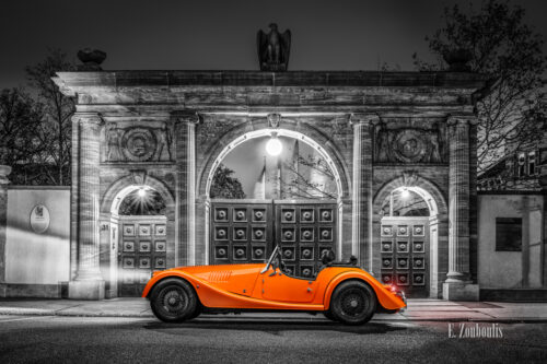 Schwarzweißaufnahme eines orangenen Morgan 4/4 Cabrio mit offenem Verdeck. Fahrzeug in Orange in der Mitte des Bildes. Im Hintergrund sieht man die Tore der Robert Bosch Stiftung in Stuttgart