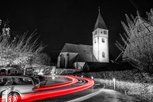 Nachtaufnahme in Nufringen an der Pelagiuskirche bei Nacht. Schwarzweiß Aufnahme mit Roten Lichtspuren, die den vorbeifahrenden Verkehr kennzeichnen