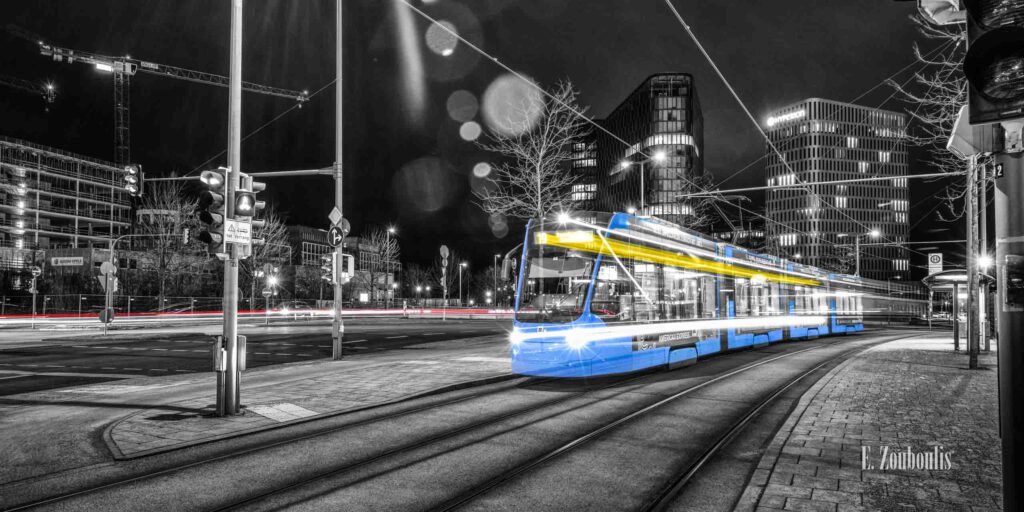 Eine Tram in München, die in die Haltestelle Vogelweideplatz einfährt. Schwarz-weiss Fotografie mit den selektiven Farben Blau, Geld und Rot, die die einfahrende Straßenbahn und den Verkehr im Hintergrund kennzeichnen.